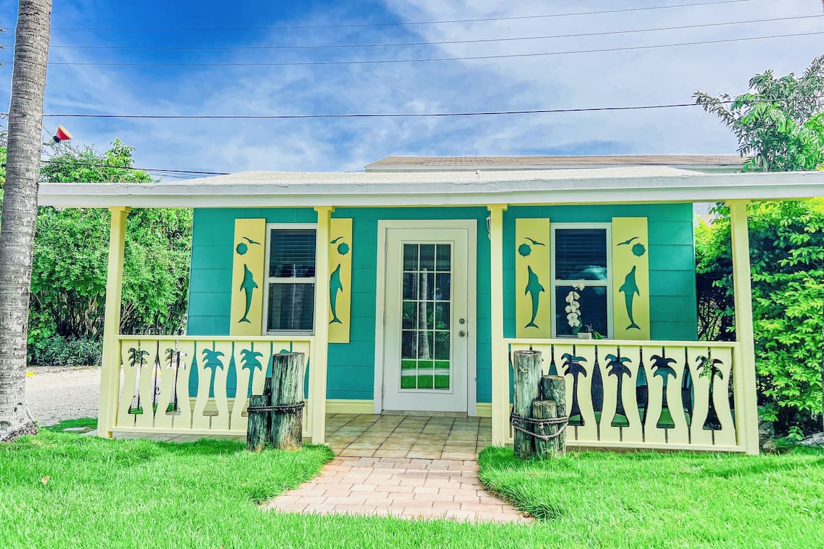 A two-story house with a green and yellow exterior, a front porch with a railing, and a pathway leading to the door.