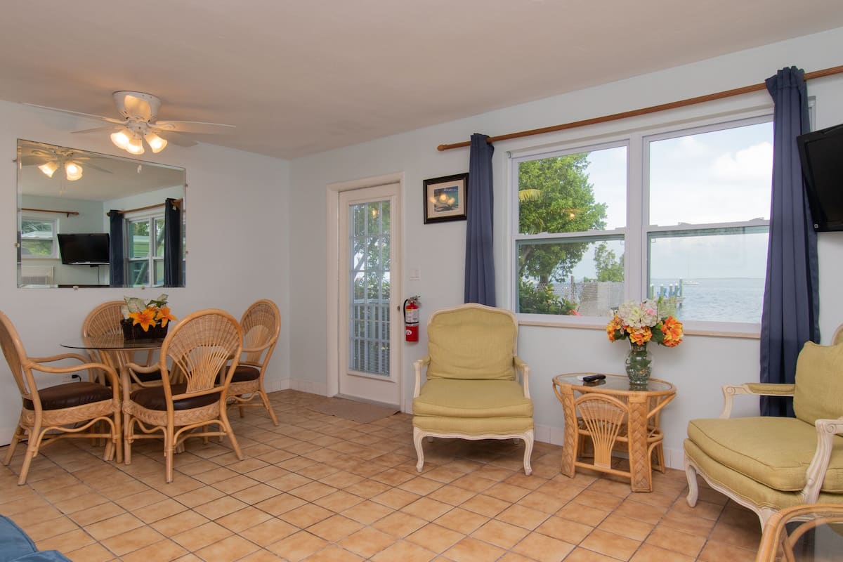 A room with a dining table, wicker chairs, a yellow armchair, a small side table with a vase, and a large window with curtains.