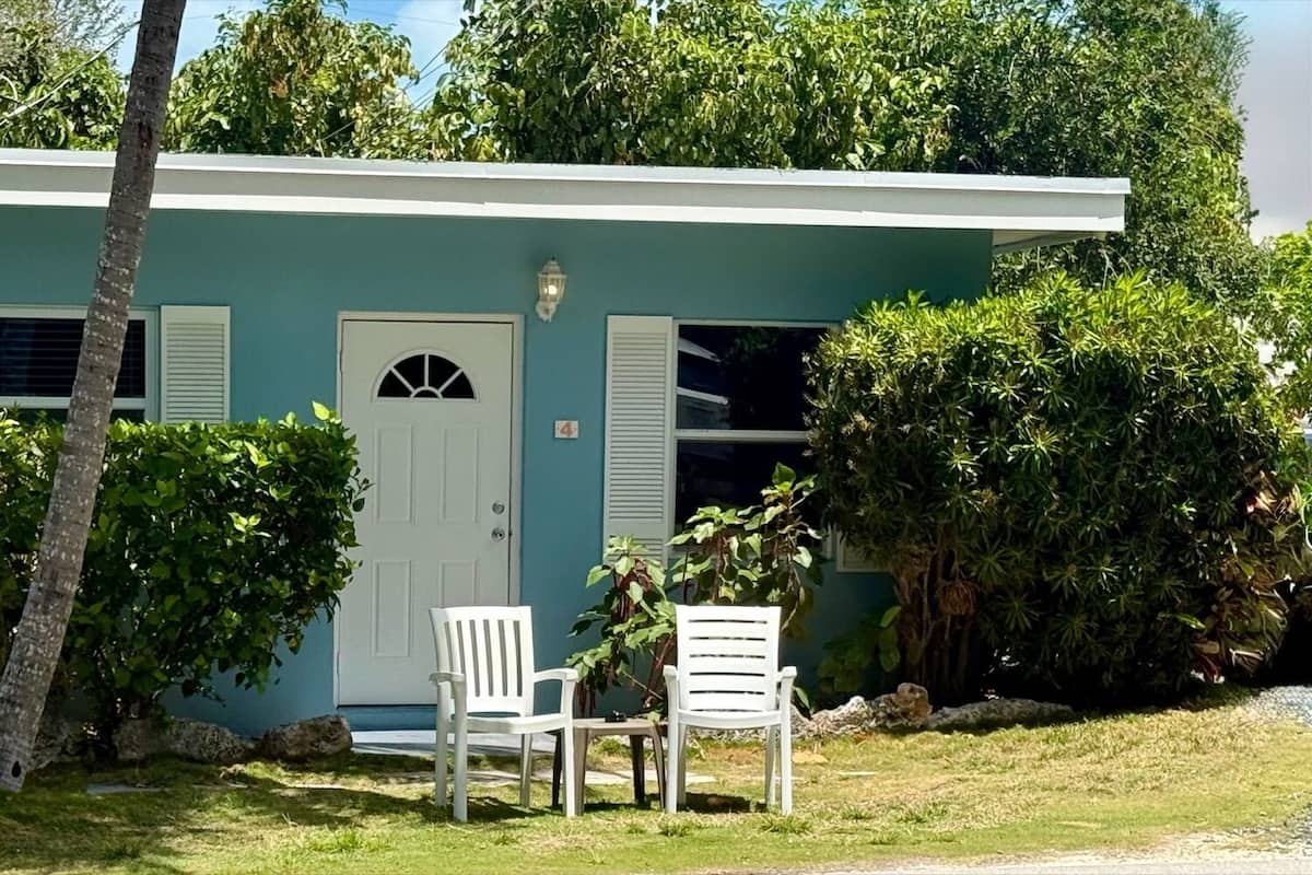 A single-story building with a white door, two windows with shutters, and a small porch with two white chairs.
