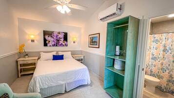 Ein Hotelzimmer mit Bett, Nachttisch, Wandairconditioner, Deckenventilator, Badezimmer mit Duschvorhang und einer grünen Regalwand.