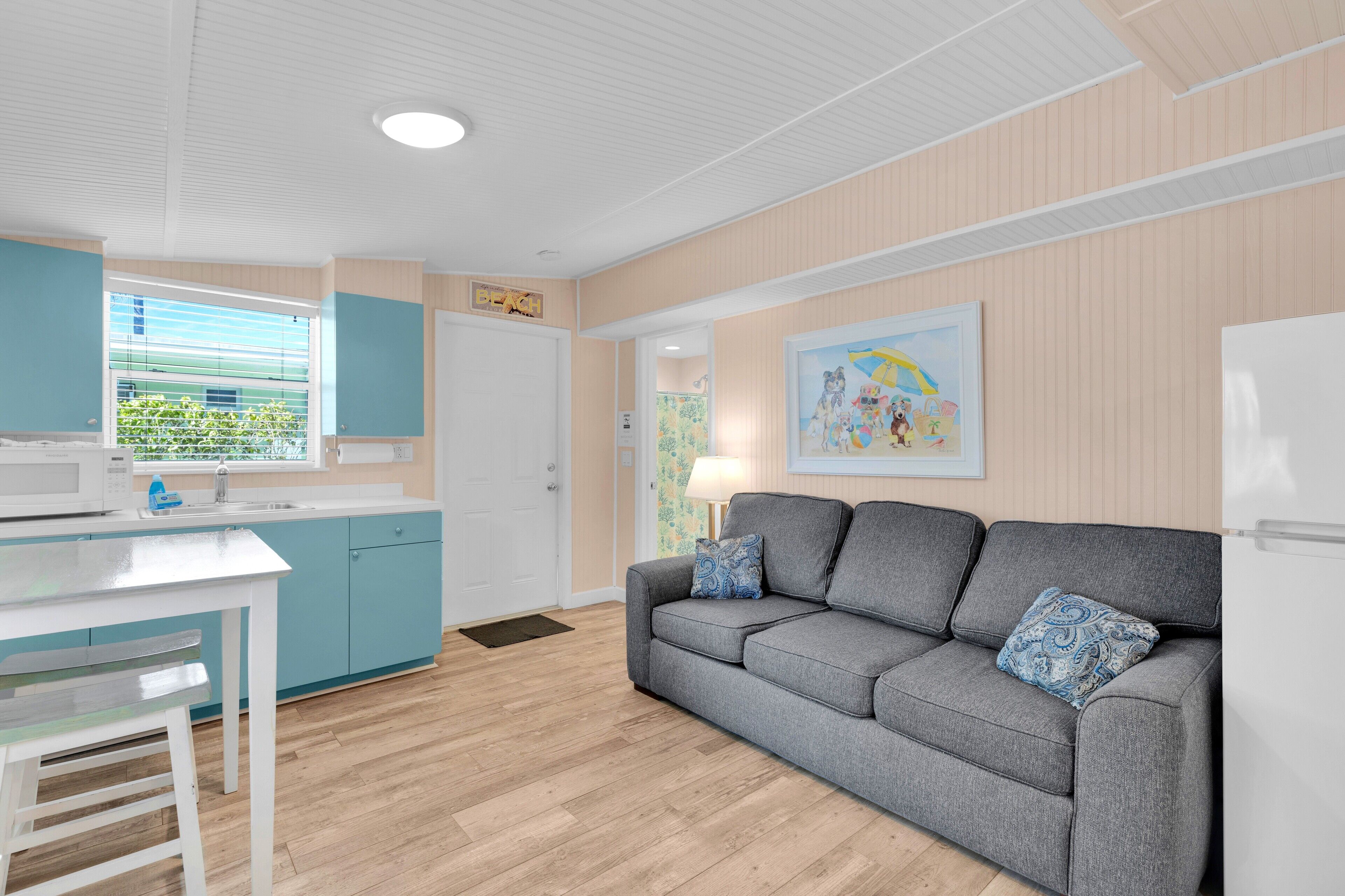 A modern kitchen with a white countertop and blue cabinets, a grey sofa with patterned cushions, and a wooden floor.
