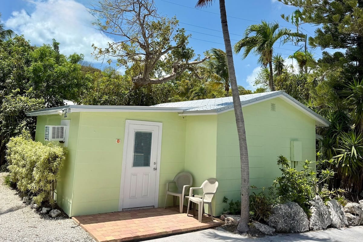 A small, single-story building with a white door, a small porch area with two chairs, and a palm tree in front.