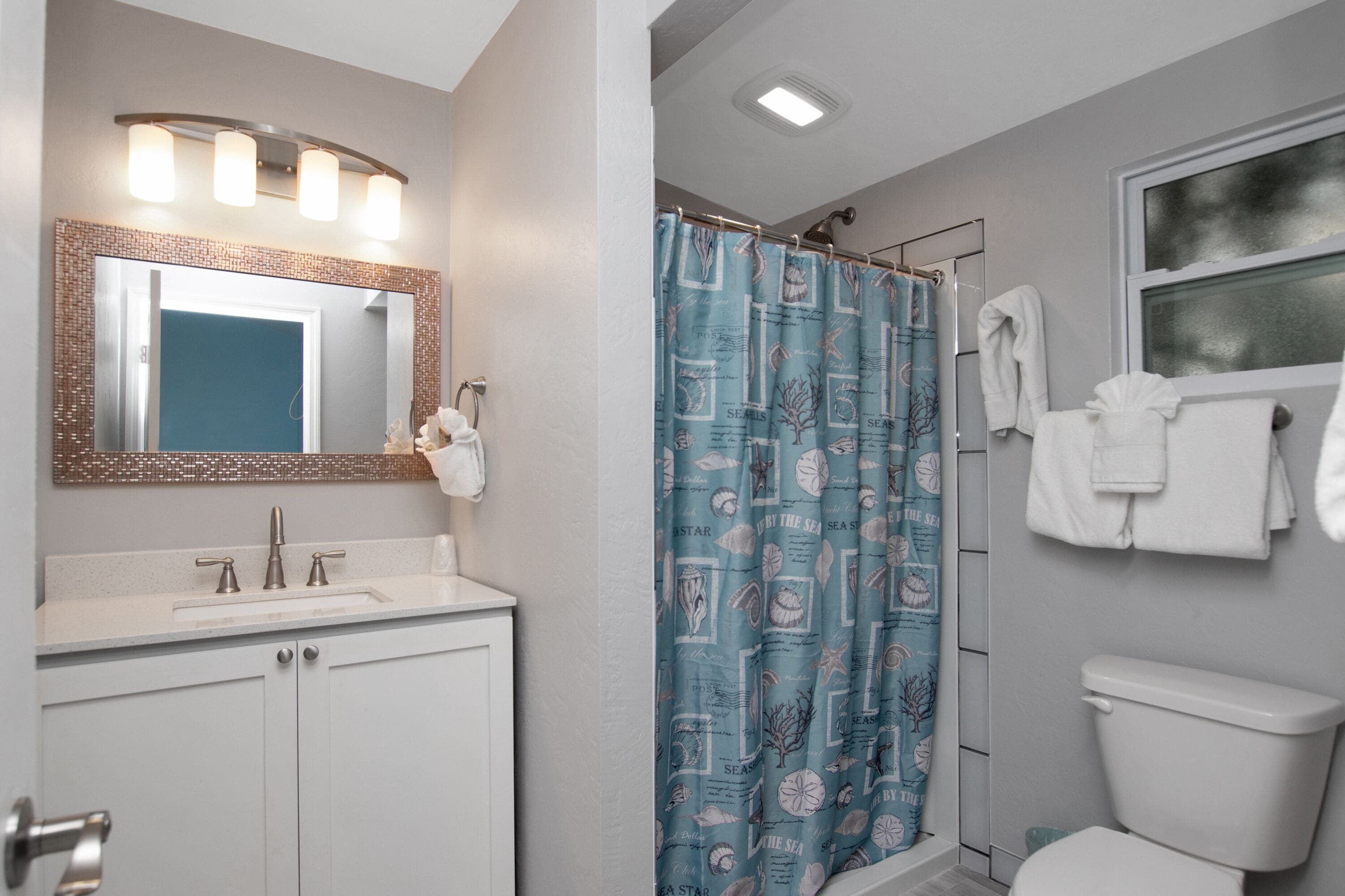 A bathroom with a white sink, a mirror with lights, a shower curtain with a sea theme, and a toilet.