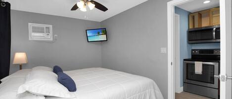 A bedroom with a bed, a ceiling fan, a TV, and a kitchen area with an oven and microwave.