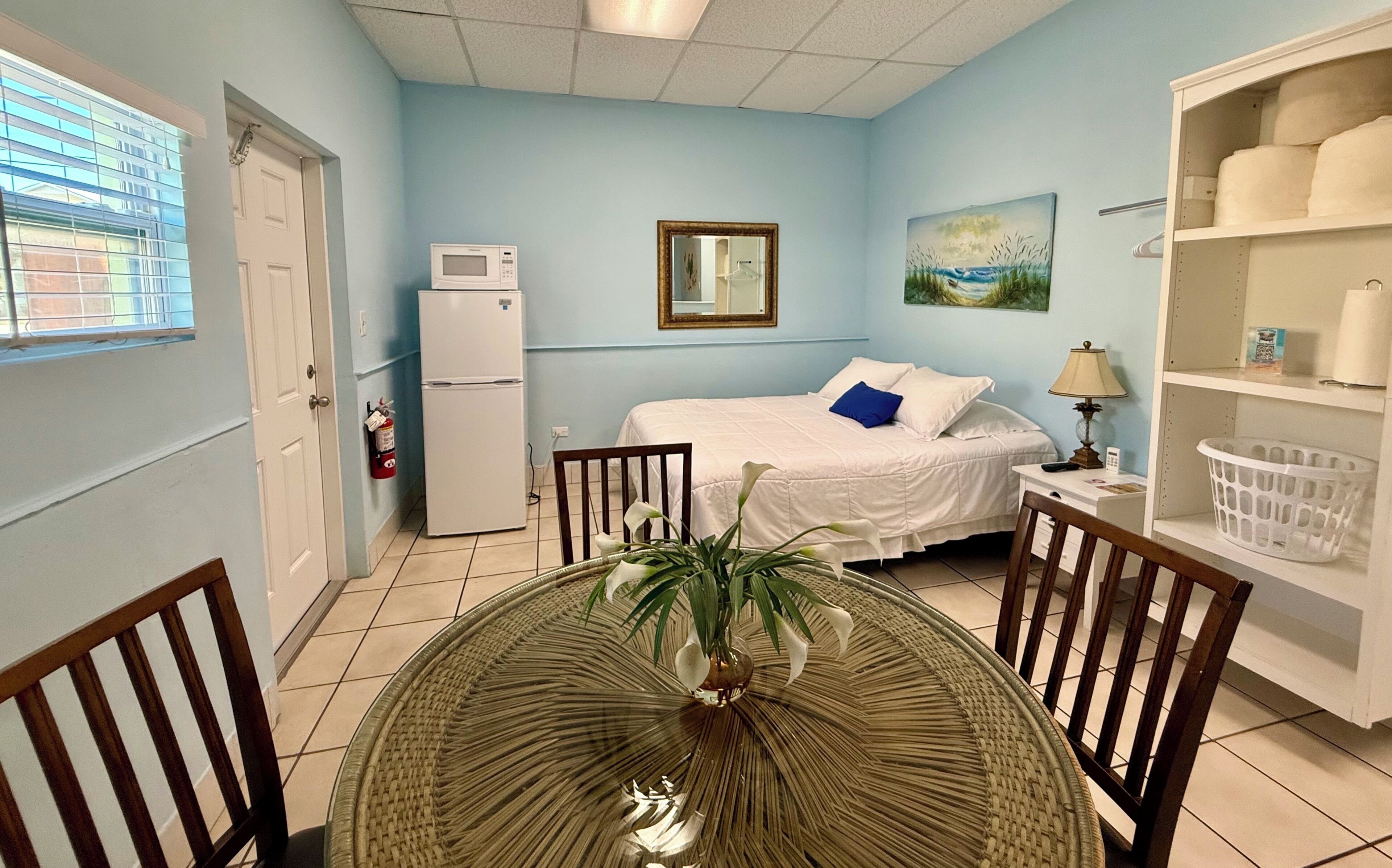 A room with a bed, a microwave, a painting, a lamp, and a basket.