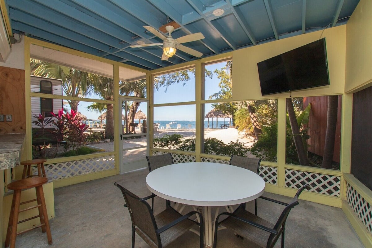 A covered outdoor dining area with a table and chairs, a ceiling fan, and a view of a beach and palm trees.