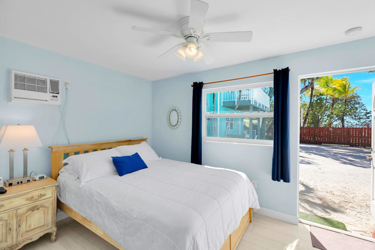 A bedroom with a bed, a nightstand, a ceiling fan, a window with curtains, and a view of the outside.