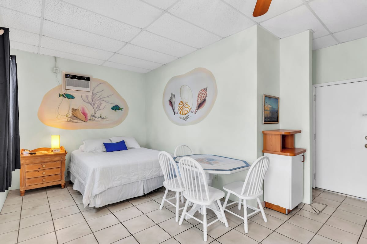 A room with a bed, a desk with chairs, a small cabinet, and a wall with a mural.