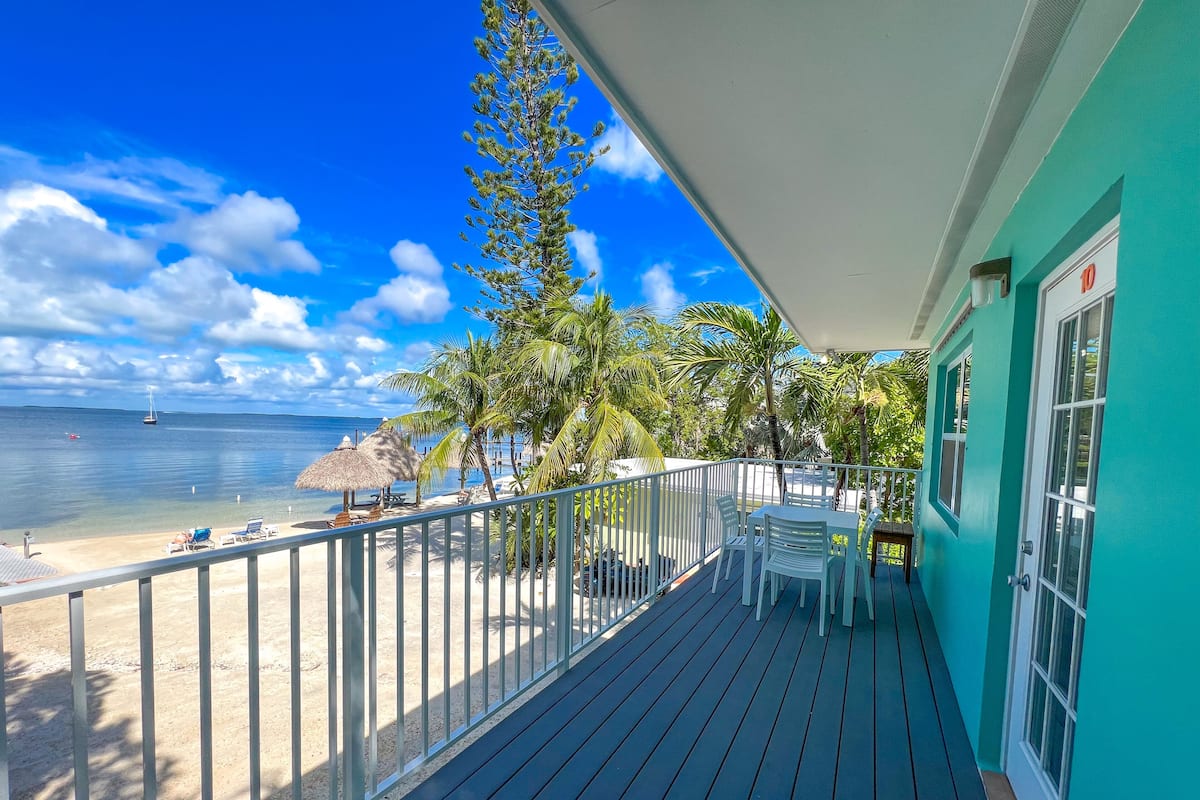 A balcony with a view of the beach, a table with chairs, and a thatched umbrella.