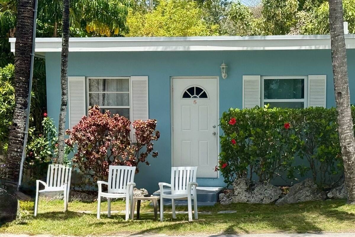 A single-story building with a white door, two windows with shutters, and a small porch with white chairs.