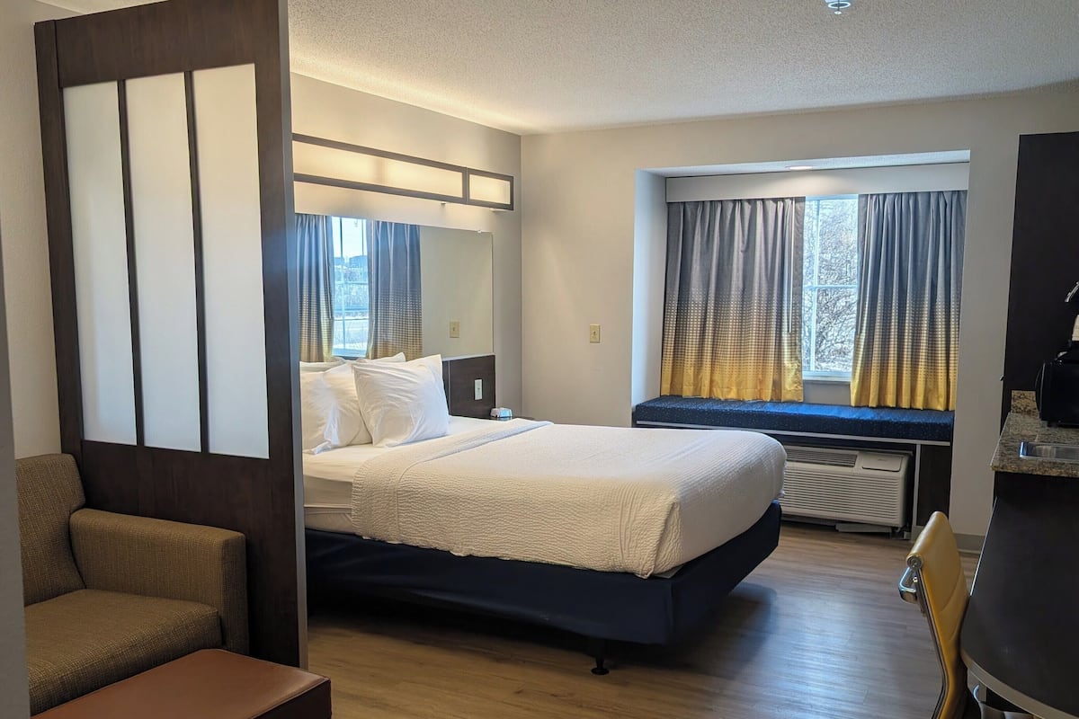 A hotel room with a bed, a seating area with a chair and a bench, a window with curtains, and a kitchenette area.