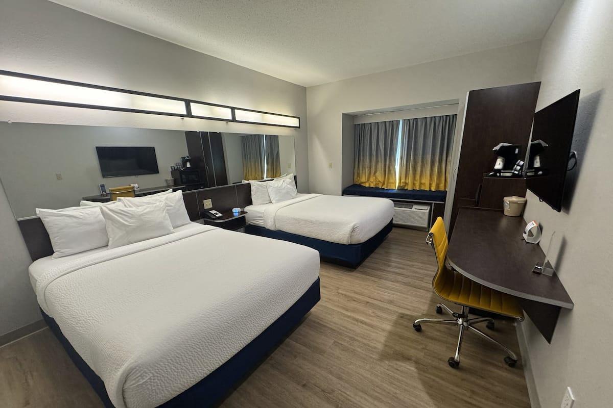 A hotel room with two beds, a desk, a chair, a mirror, a TV, and a window with curtains.