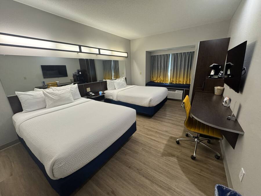 A hotel room with two beds, a desk, a chair, a mirror, a TV, and a window with curtains.
