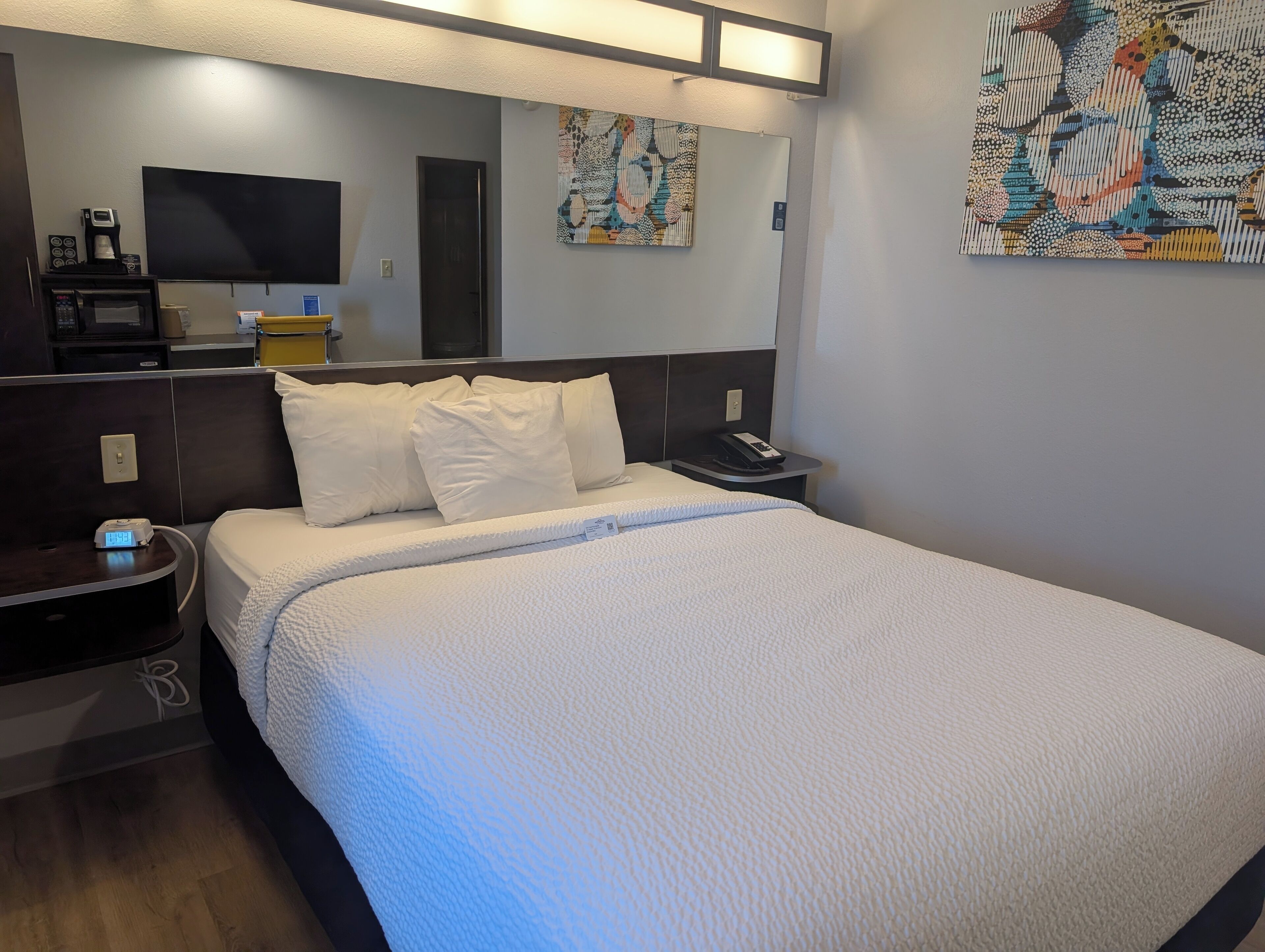 A hotel room with a bed, bedside table, a TV, a coffee machine, and artwork on the walls.
