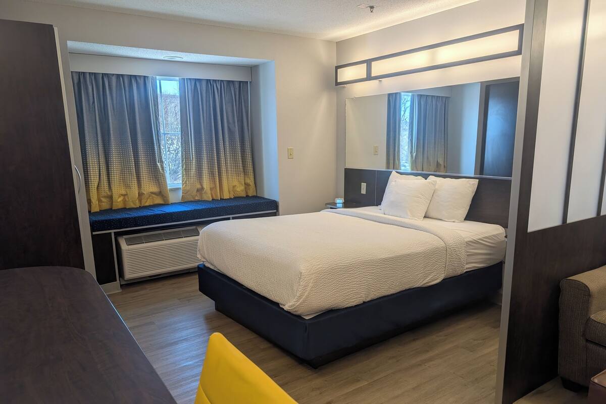 A modern hotel room with a large bed, a bedside table, a chair, and a window with curtains.