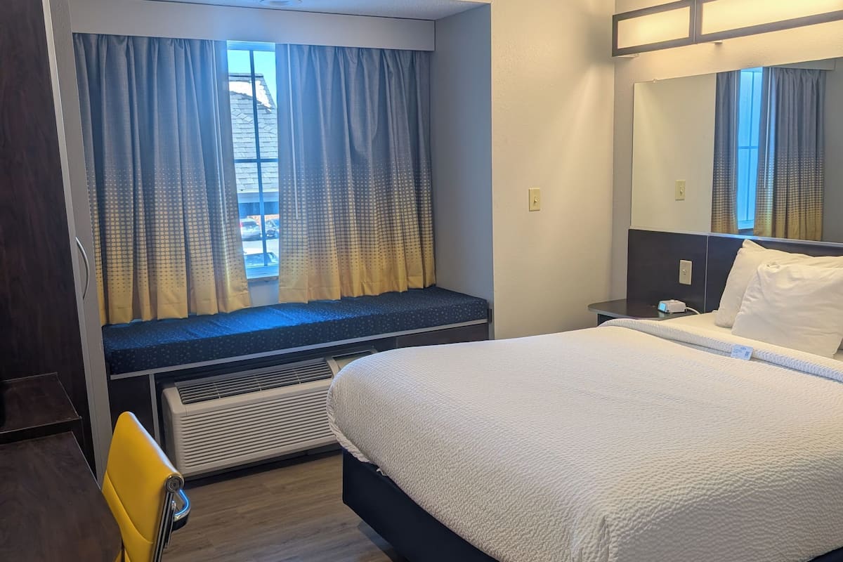 A hotel room with a bed, a desk, a chair, a window with curtains, and a mirror.