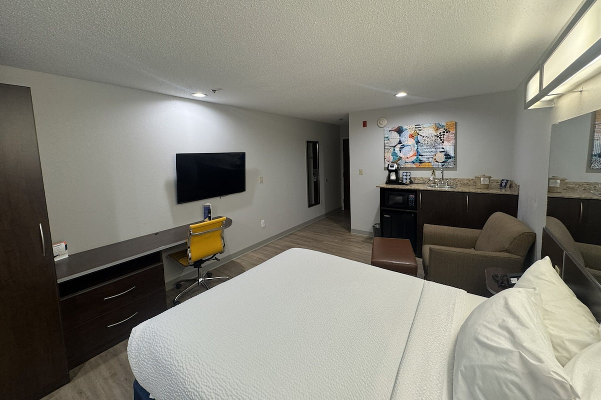 A hotel room with a bed, a desk, a chair, a TV, a kitchenette, and a bathroom.