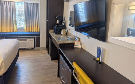 A compact hotel room with a bed, a kitchenette, a flat-screen TV, and a large mirror.
