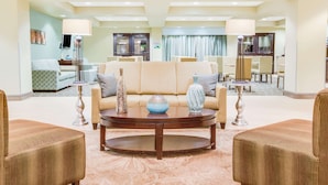 A spacious lobby with a beige sofa, two armchairs, and a coffee table. There are two lamps, a television, and a dining area in the background. - Wingate by Wyndham Bossier City, LA (Bossier City)