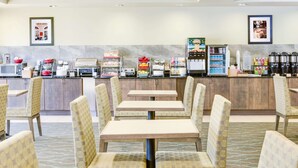 A hotel dining area with multiple tables and chairs, a self-service beverage station, and a kitchen area in the background. - Wingate by Wyndham Bossier City, LA (Bossier City)