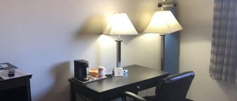 Hotellihuone, jossa on työpöytä, kaksi lamppua, tuoli, kahvinkeitin ja sänky.