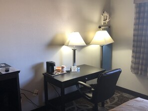A hotel room with a desk, two lamps, a chair, a coffee maker, and a bed.