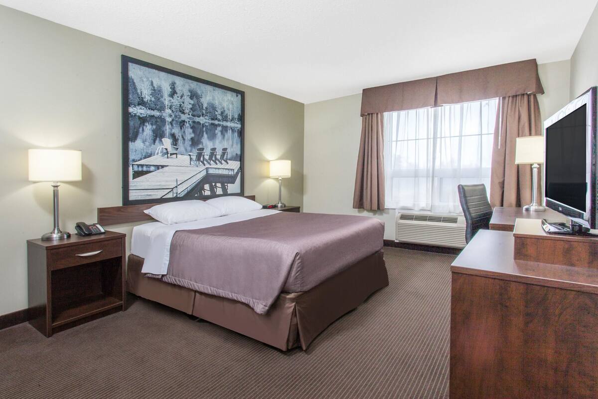 Ein Hotelzimmer mit einem großen Bett, zwei Nachttischen mit Lampen, einem Schreibtisch mit Fernseher und einem Gemälde von einem Steg und See.