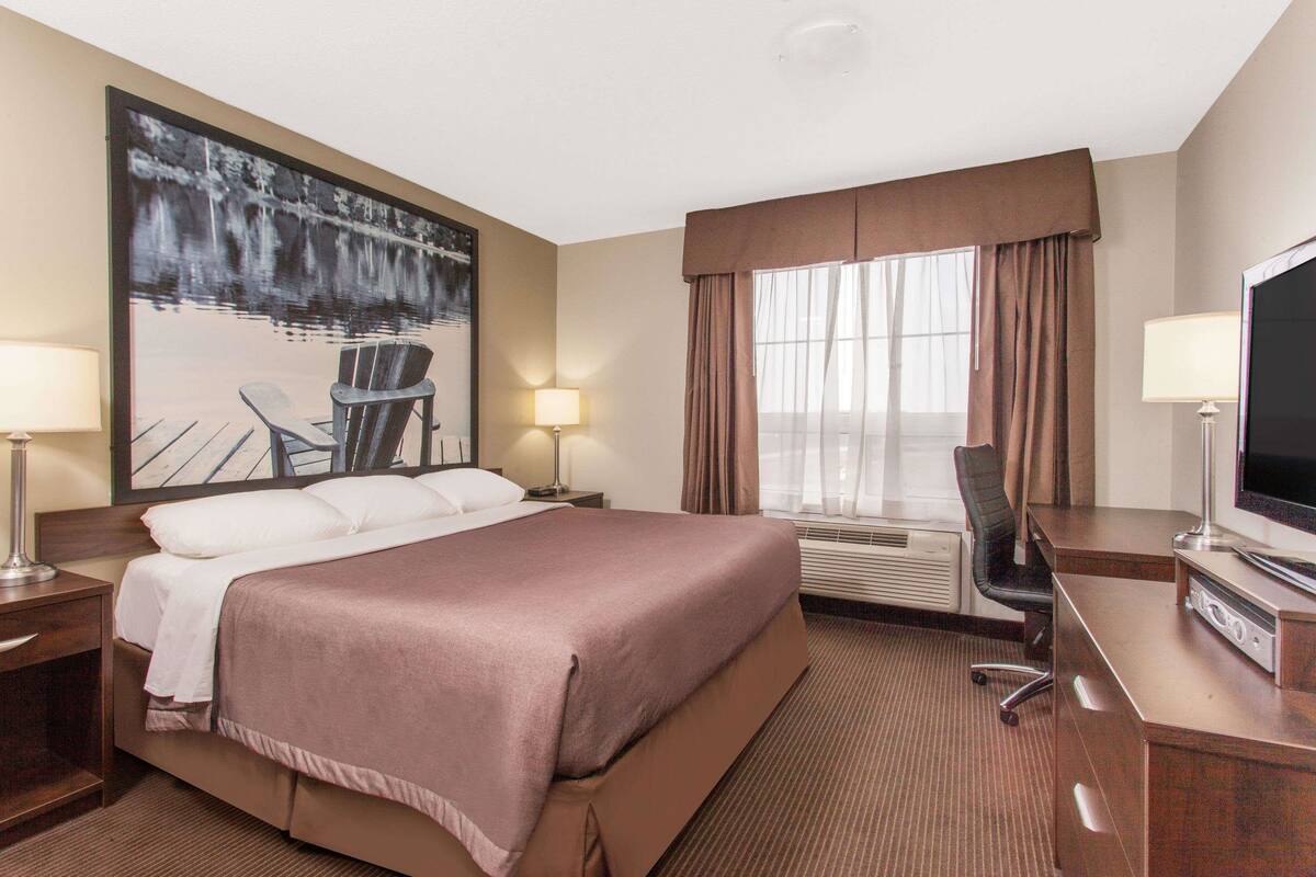 Ein Hotelzimmer mit einem großen Bett, einem Schreibtisch mit Computer, einem Fernseher und einem Gemälde eines Sees.