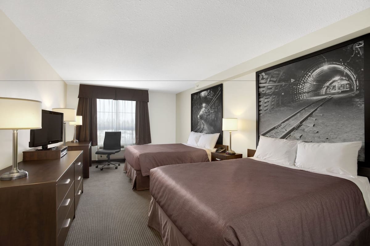 Ein Hotelzimmer mit zwei Betten, einem Schreibtisch mit Fernseher, einem Sessel, einer Schreibtischlampe, einem Fenster mit Vorhängen und einem großen Schwarz-Weiß-Foto an der Wand.