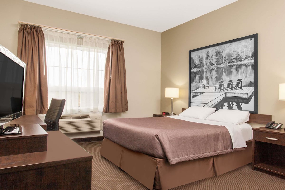 Ein Hotelzimmer mit einem großen Bett, einem Schreibtisch mit Stuhl, einem Fernseher, einem Fenster mit Vorhängen, einer Lampe und einem Bild mit Seeblick.