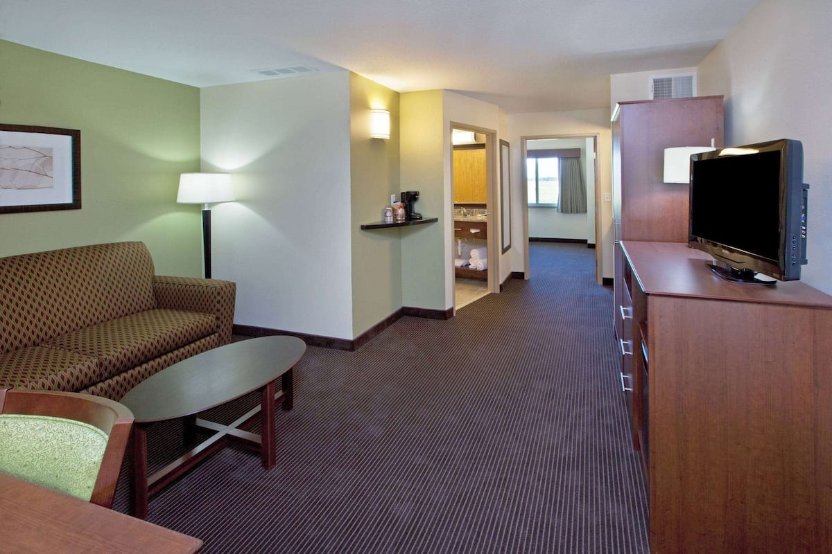 Une chambre d’hôtel avec un canapé, une petite table, un téléviseur posé sur un meuble en bois, et un couloir menant à la salle de bain.