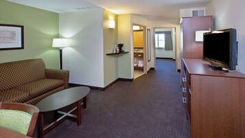 A hotel room with a sofa, a small table, a TV on a wooden cabinet, and a hallway leading to a bathroom.