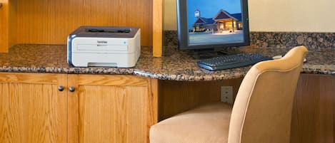 Escritorio de madera con una impresora Brother, un monitor de computadora mostrando una escena de hotel y una silla color beige.