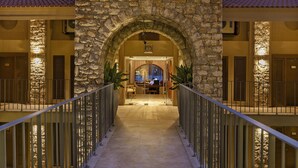 Un arco de piedra que conduce a una zona de recepción con un mostrador y sillas, flanqueado por barandillas y plantas en macetas.