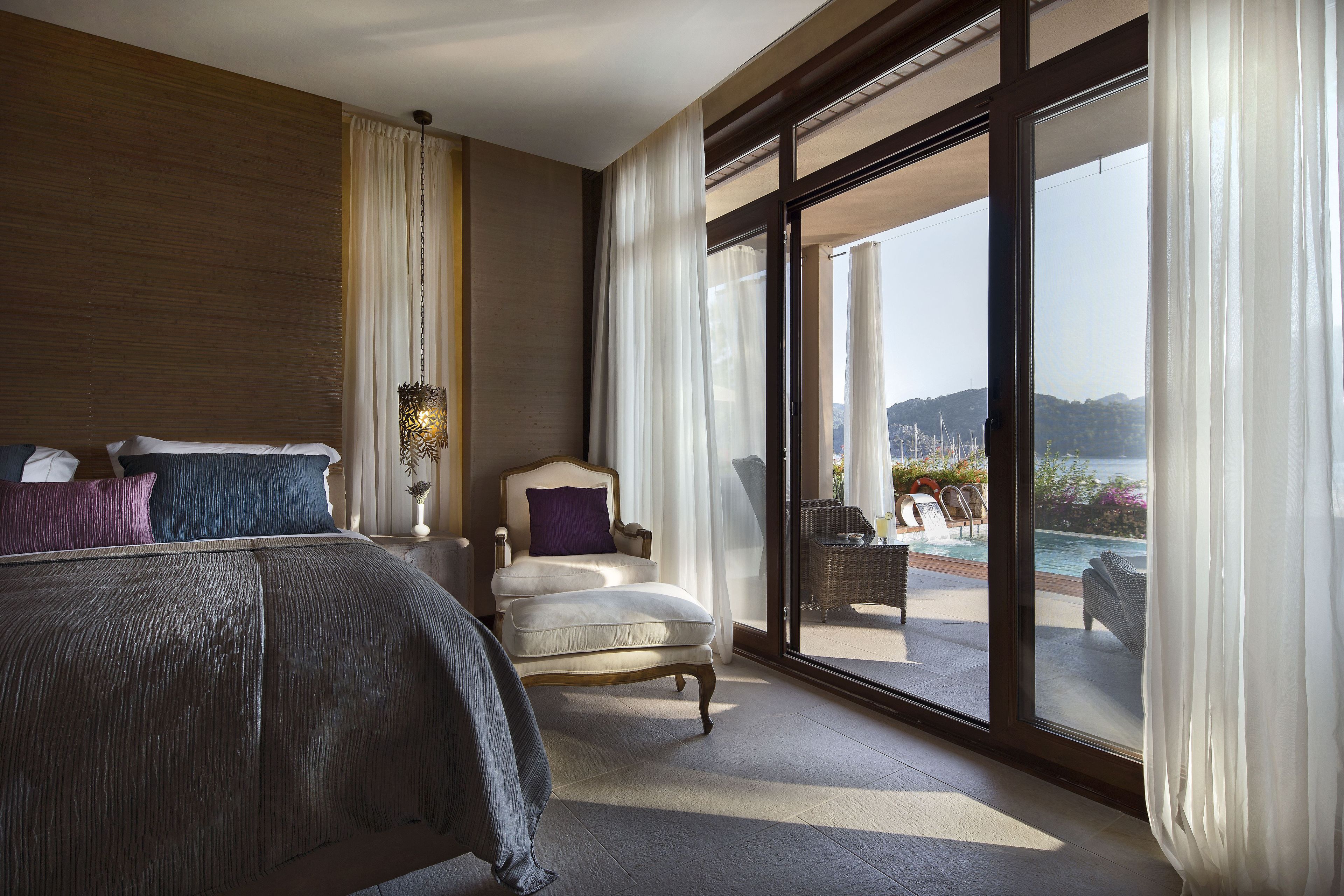 A spacious bedroom with a large bed, a chair, and a view of a pool and mountains through floor-to-ceiling windows.