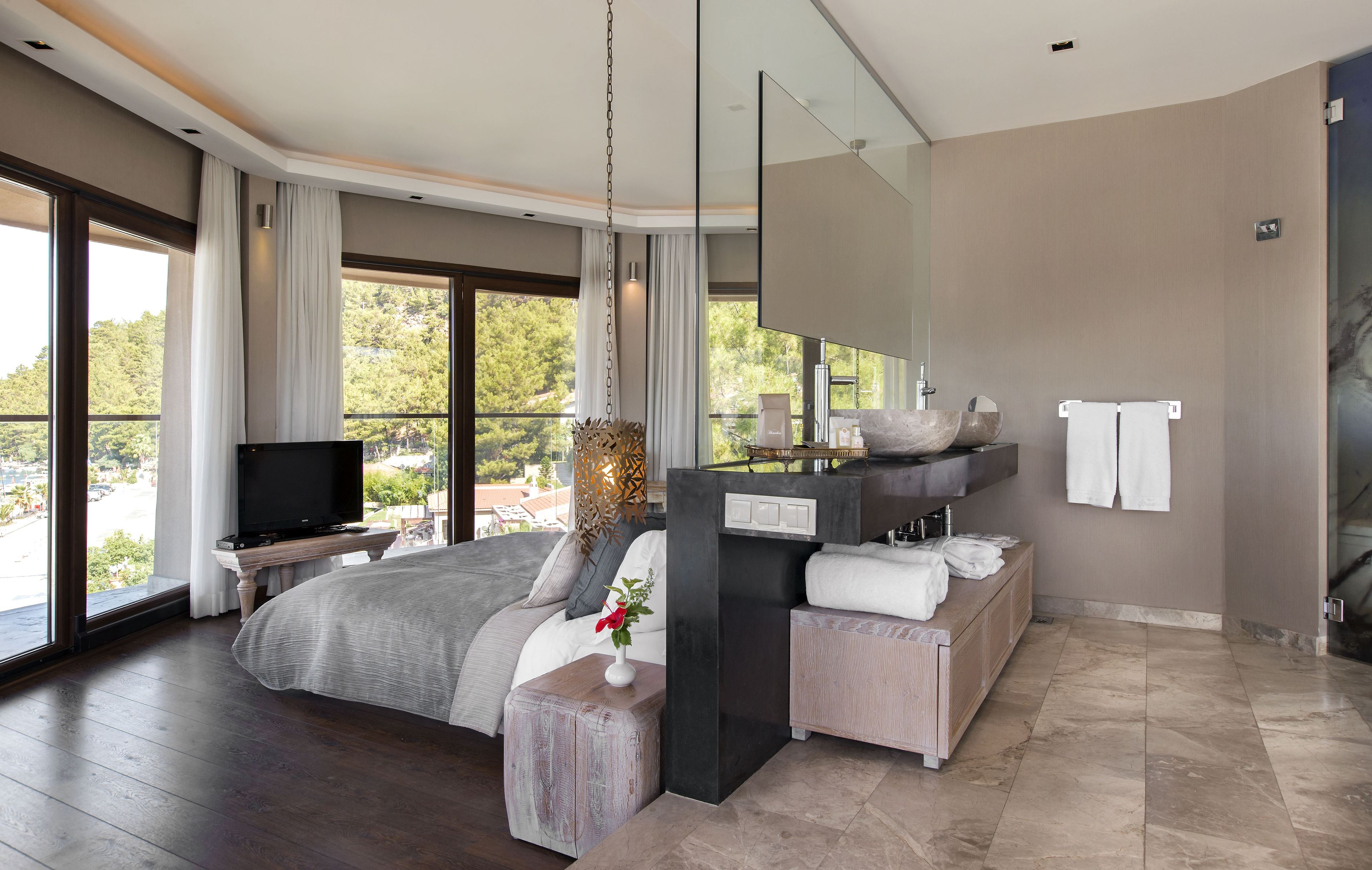 A modern bedroom with a large bed, a TV, a bathroom with a sink and mirror, and a view of the outdoors.