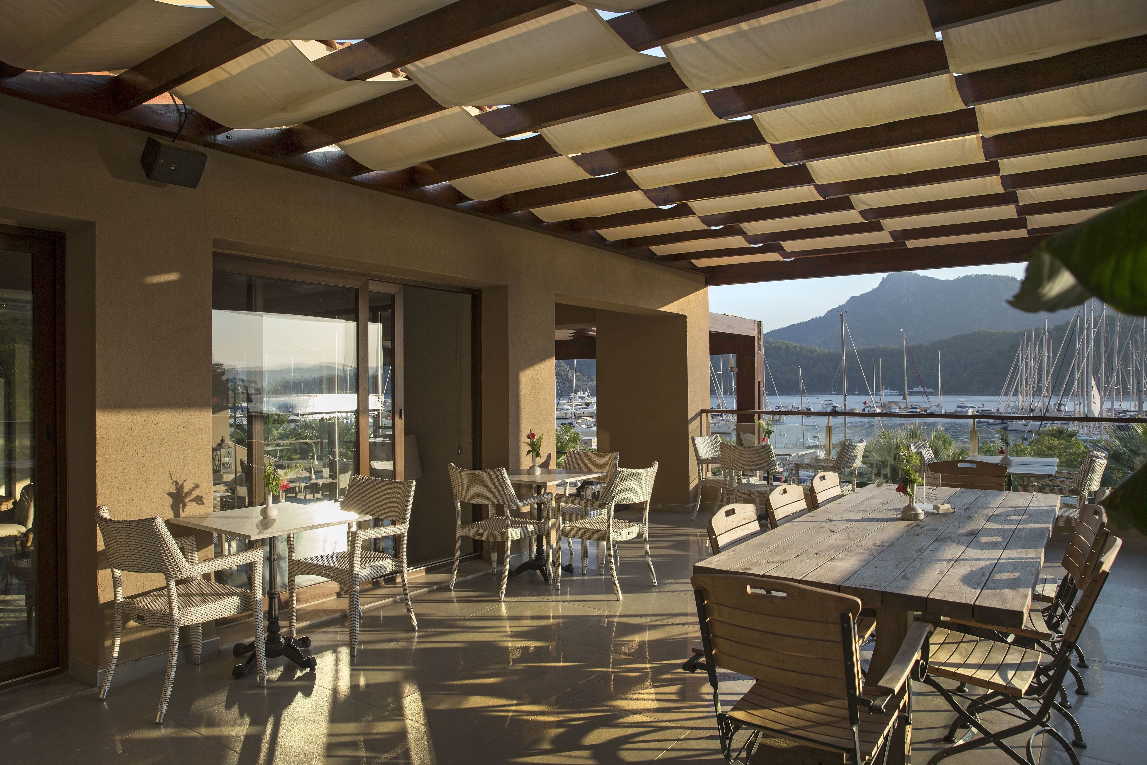 An outdoor patio with tables and chairs, overlooking a marina with boats and a mountainous backdrop.