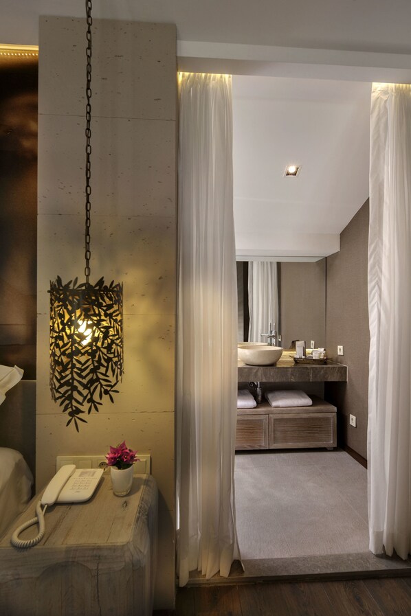 A modern bathroom with a large mirror, a sink, and a toilet. There is a telephone and a vase with flowers on a table. - Martı Hemithea Hotel (Marmaris)