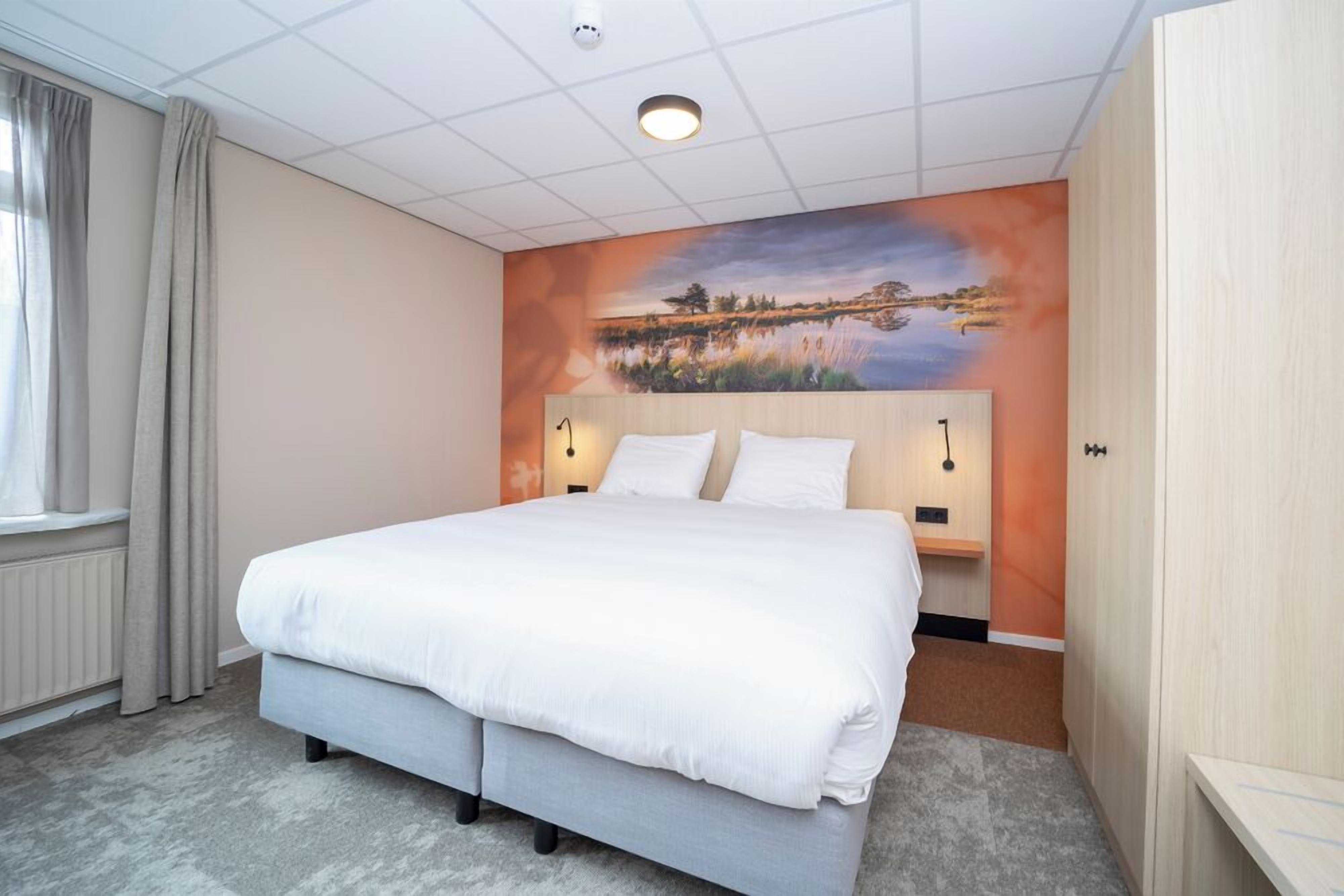 A hotel room with a large bed, a painting on the wall, and a window with curtains.