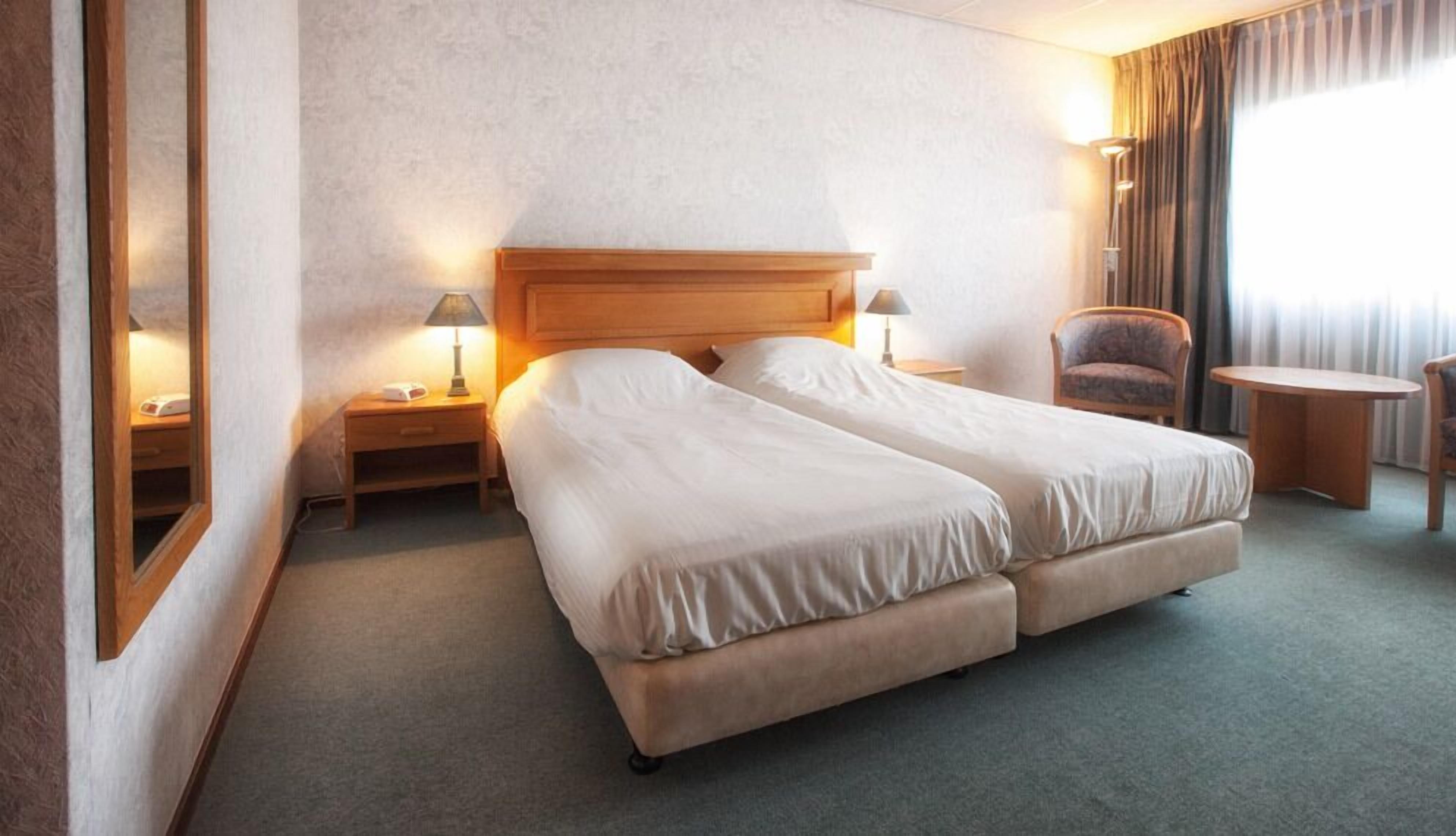 Habitación de hotel con una cama doble, dos mesitas de noche con lámparas, una silla y una pequeña mesa.