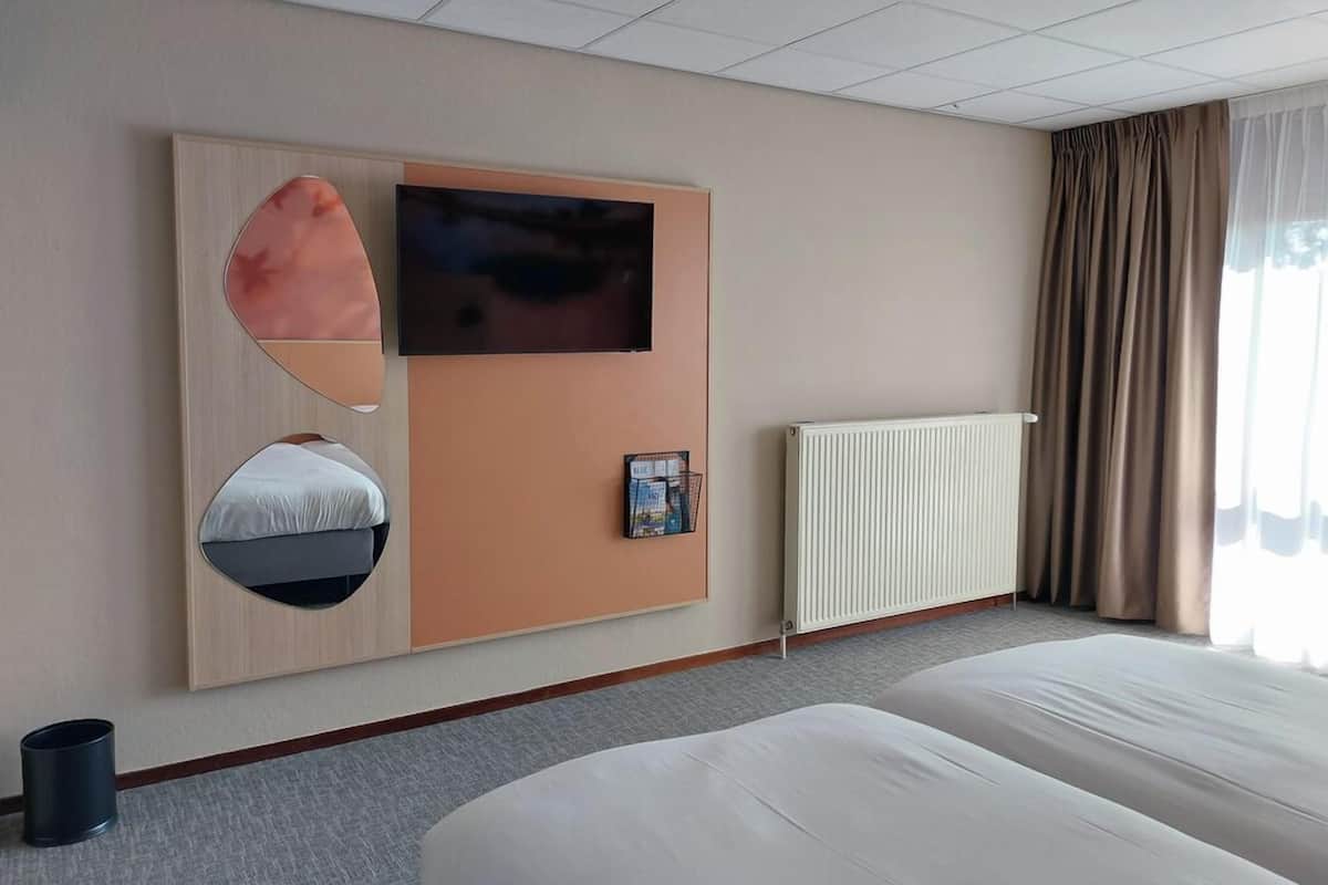 Ein Hotelzimmer mit einem großen Bett, einem an der Wand befestigten Fernseher, einem Spiegel, einem Zeitschriftenständer und einer Heizung.