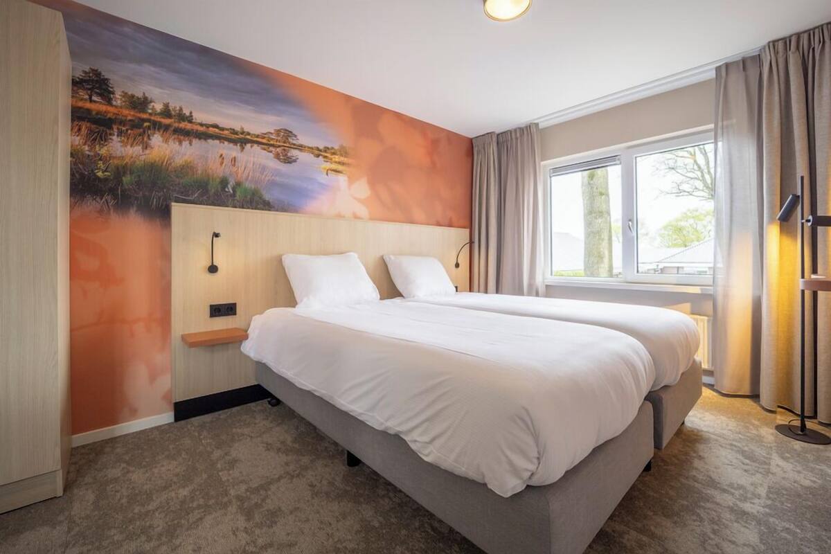 Ein Hotelzimmer mit zwei Betten, einem großen Fenster, einem Gemälde an der Wand und einem Nachttisch mit einer Lampe.