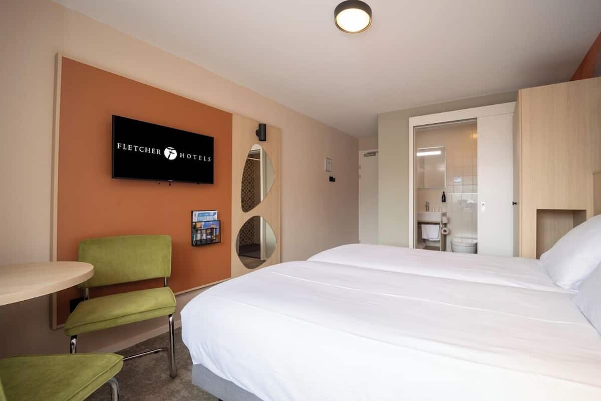 Ein Hotelzimmer mit Bett, Wandfernseher, grünem Sessel und kleinem Tisch.
