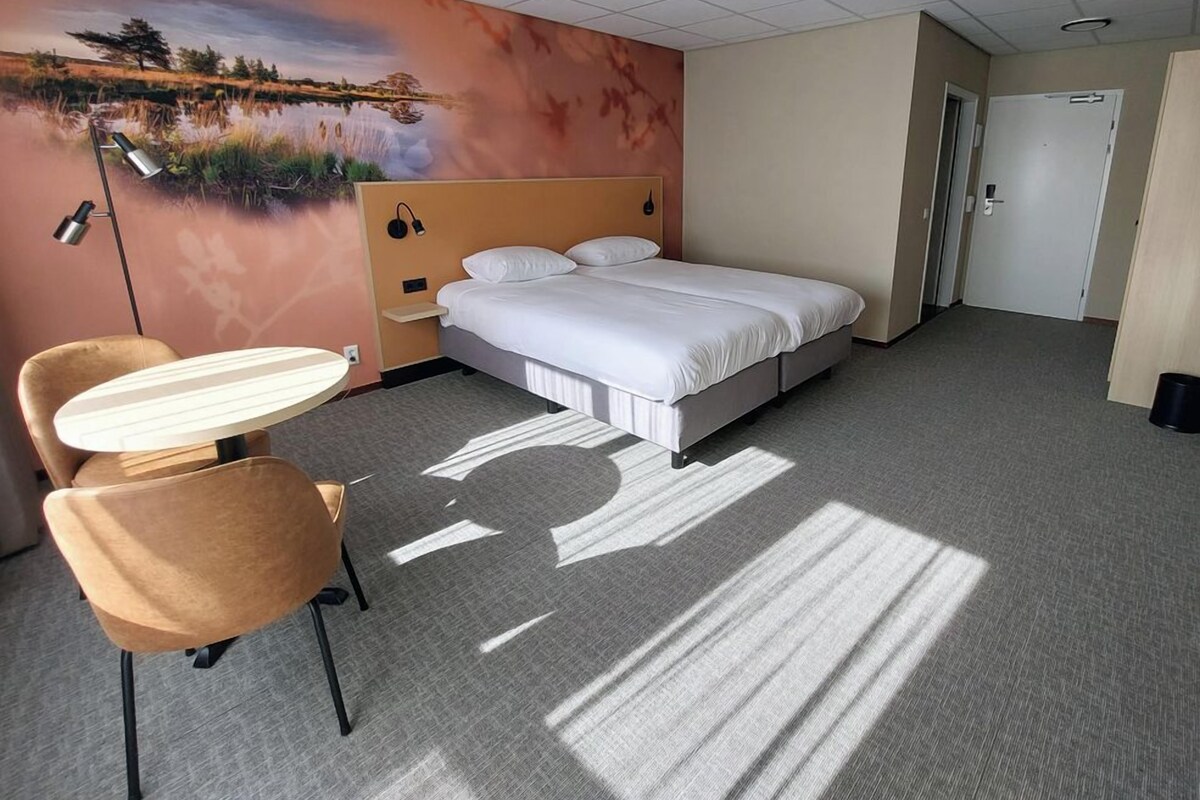 Una habitación de hotel con cama, escritorio, silla, un mural en la pared y una puerta.