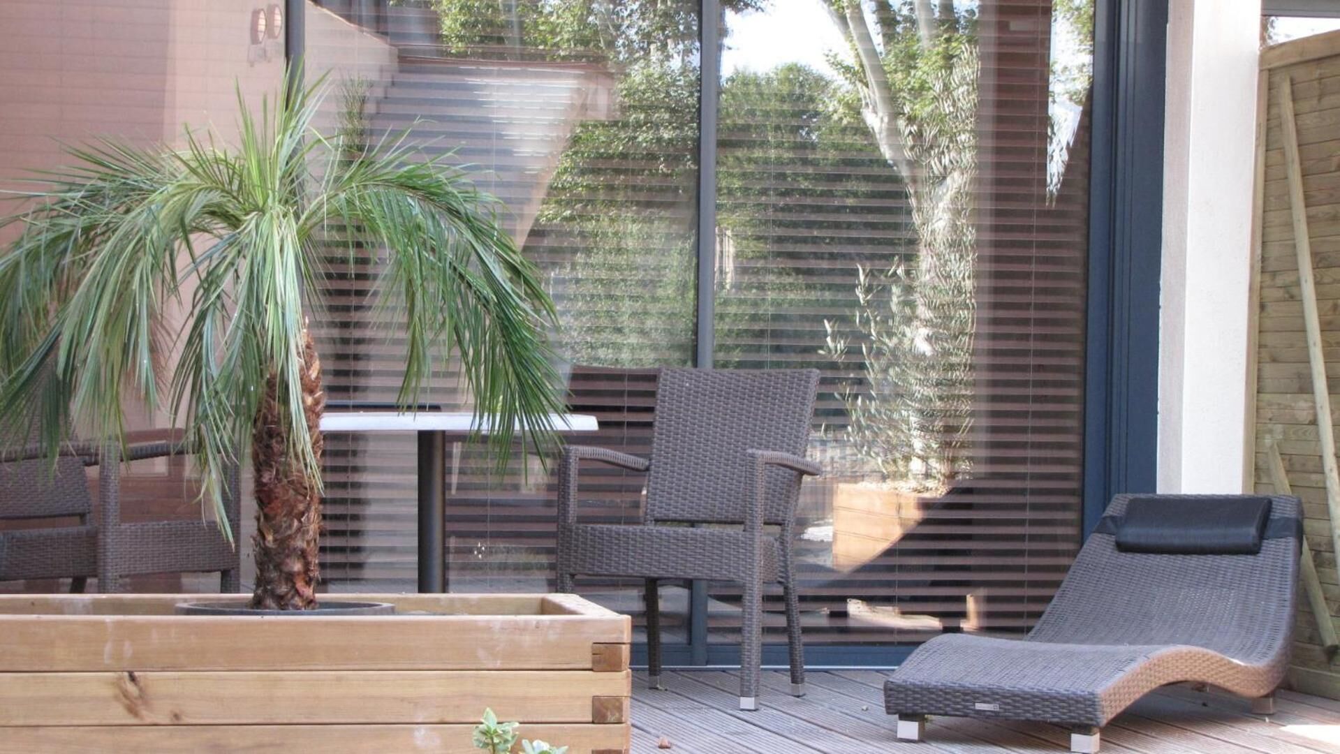 Eine Terrasse mit Korbmöbeln, einem Holzpflanztopf mit einer Palme und einer Glaswand mit Blick auf ein Gebäude und Bäume.