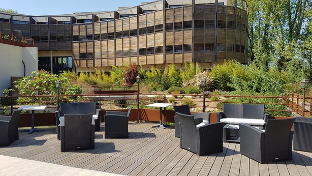 Un patio al aire libre con mobiliario de mimbre y mesas, rodeado por una terraza de madera y abundante vegetación.