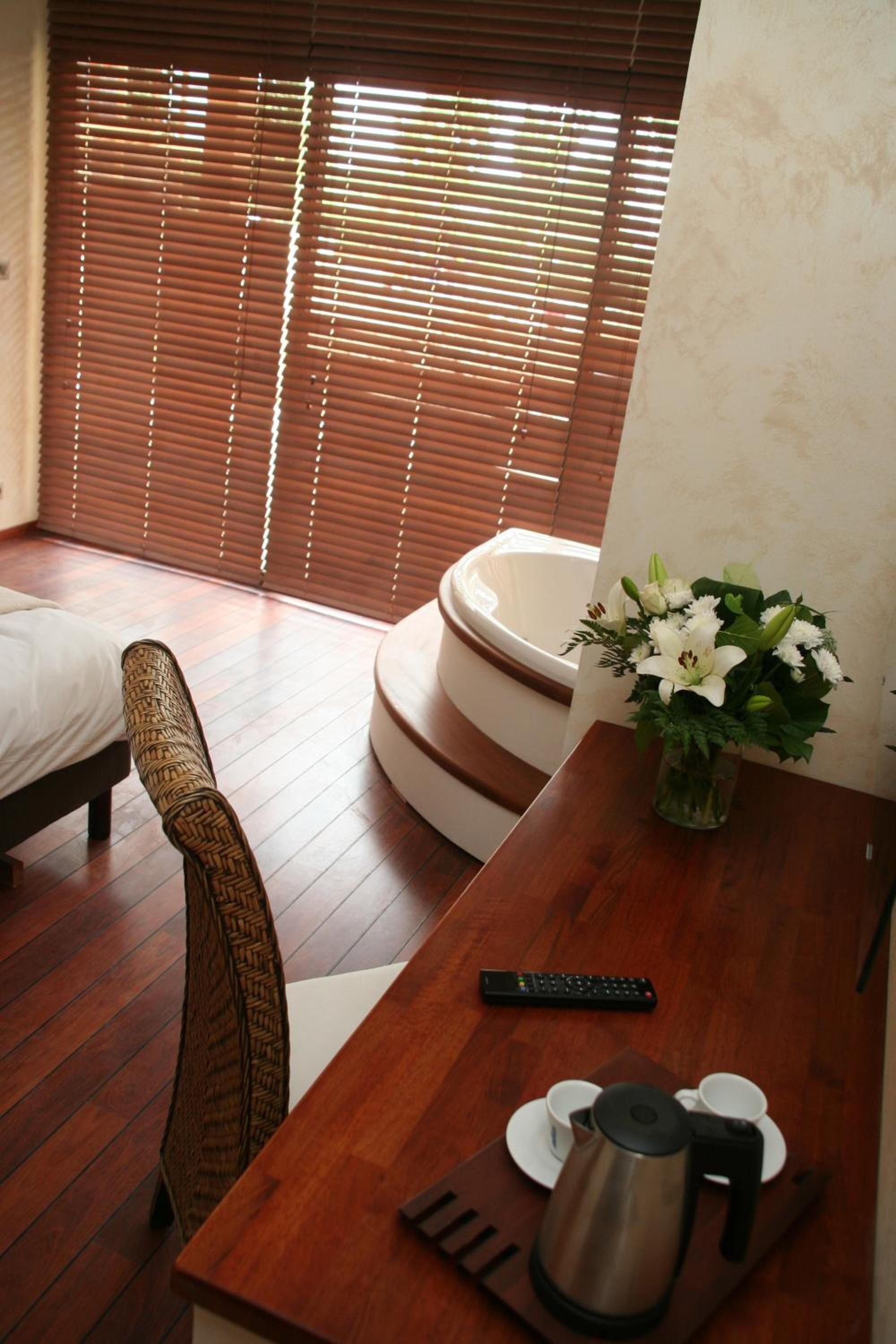 A hotel room with a wooden floor, a bed, a chair, a small table with a remote control, a coffee pot, and a vase with flowers.