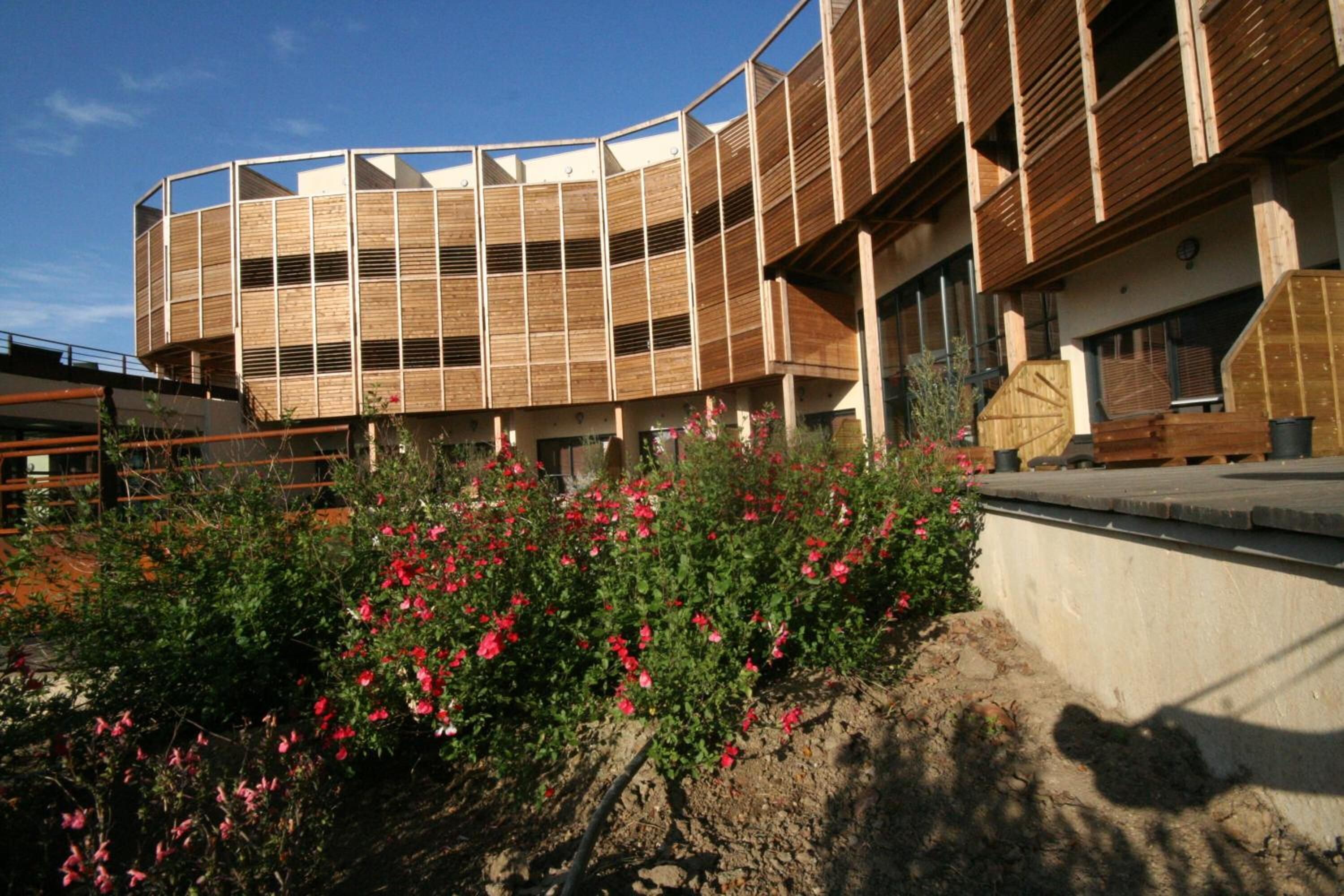 Un edificio moderno con fachada de madera curvada y un jardín con flores rojas al frente.