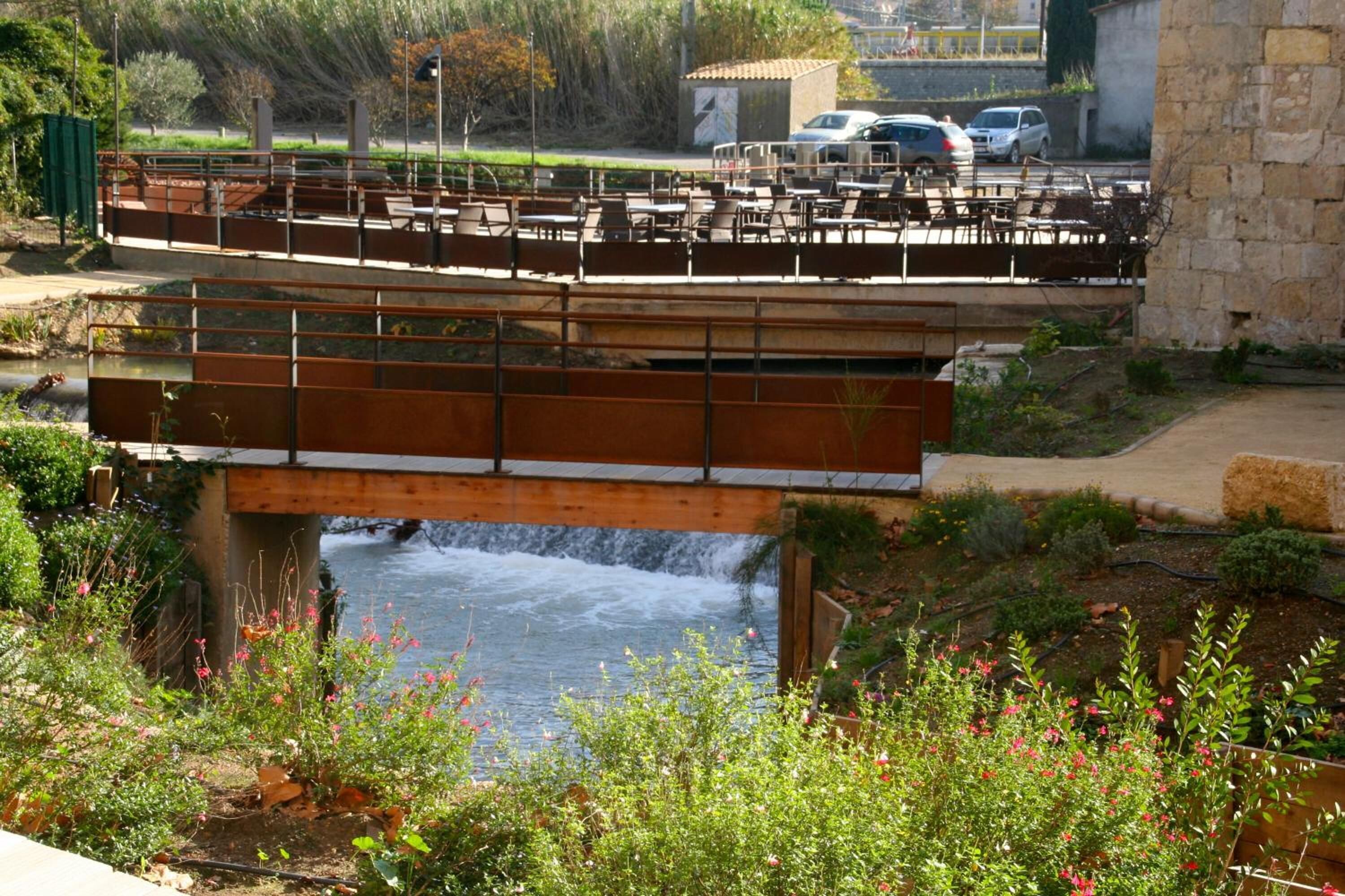 A bridge with seating overlooking a flowing river, surrounded by greenery and a stone wall.
