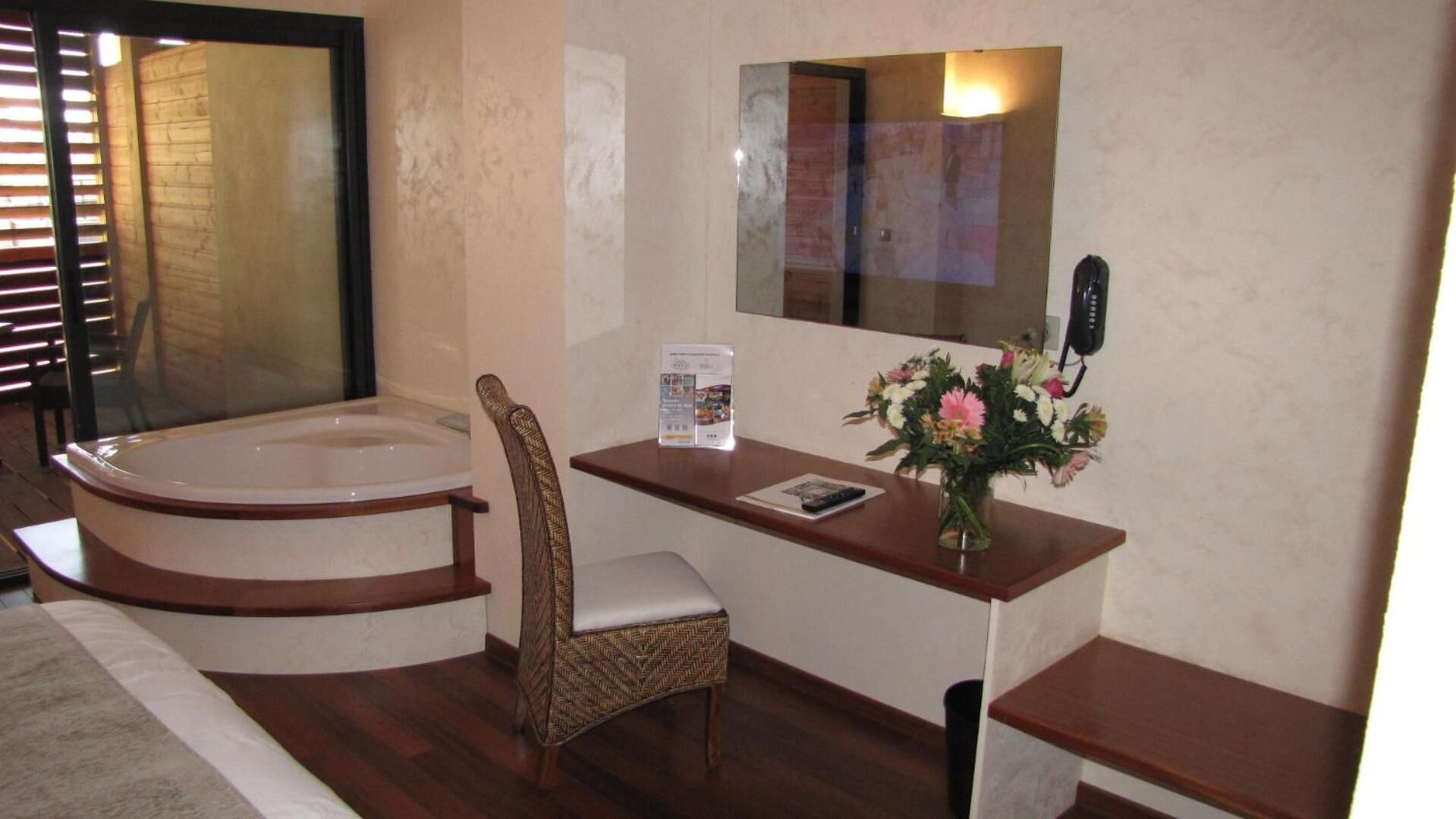 Ein Badezimmer mit einer Badewanne, einem Sessel, einem Schminktisch mit Spiegel, einem Telefon und einer Vase mit Blumen.